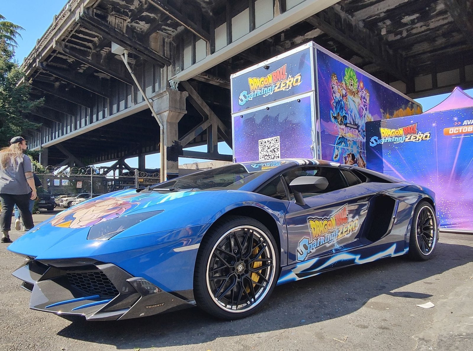 Photograph of a Lamborghini decorated with the Dragon Ball Sparking! Zero game logo on the side, and SSJ Blue Goku on the car's hood. It's parked next to a Dragon Ball Sparking! Zero trailer