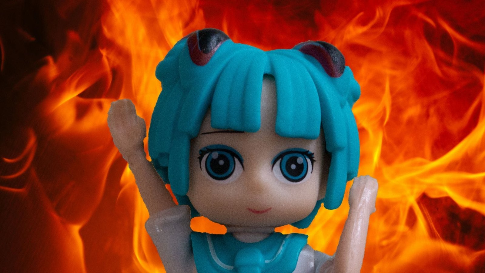 Photograph of a green-haired, anime style doll against a wall of flames. The pose elicits a similar feeling to the "Burning Elmo" meme.